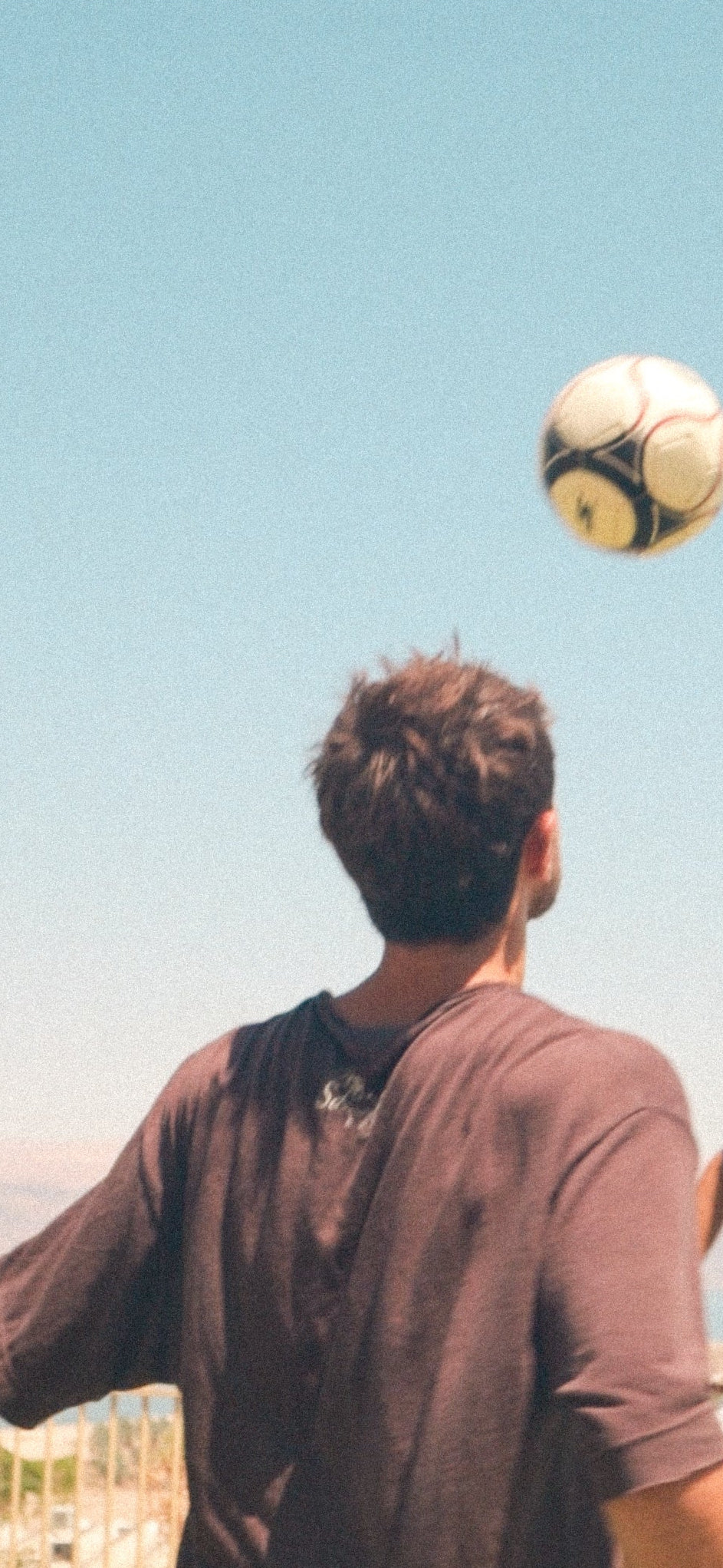 A man seen from behind wearing a dark brown t-shirt, balancing a soccer ball on his head outdoors under a clear blue sky.
